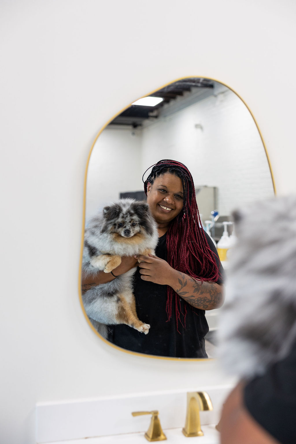 FreshCoat Grooming bather smiling in the salon mirror while holding a fluffy blue merle Pomeranian in Charlotte, NC